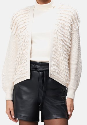 Cardigan crème texturé laine avec motifs en relief sur un col roulé blanc, associé à un short en cuir noir taille haute noué à la taille.