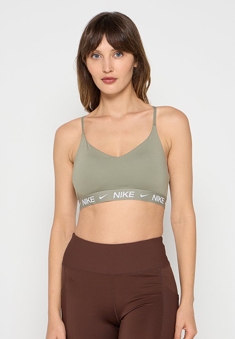 Sujetador deportivo verde oliva con tirantes finos y una cinturilla ancha gris que presenta el logo de Nike. El material parece suave con una forma ajustada.