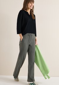 Femme marchant, portant une chemise noire à manches longues, un pantalon gris à cordon, des baskets blanches et vertes, tenant un pull vert clair plié.