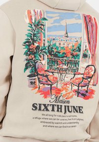 Sudadera color beige con un gráfico colorido de un balcón parisino, detalles florales, sillas y texto. Material suave y corte relajado.