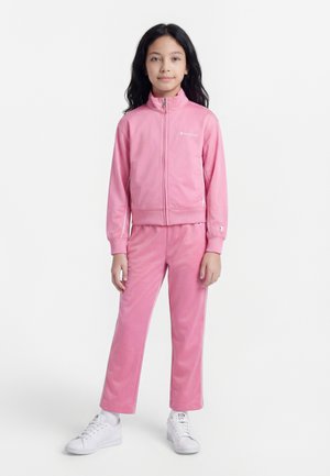 Νεαρή κοπέλα στέκεται, φορώντας ένα ροζ φόρμα με φερμουάρ και λευκά αθλητικά παπούτσια, μπροστά σε απλό λευκό φόντο.