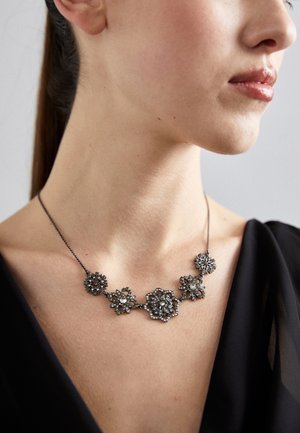 Femme portant un collier avec cinq pendentifs noirs en forme de fleur ornés de petites pierres précieuses transparentes, associé à un haut noir.