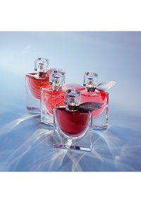 Quatre flacons de parfum en verre transparent contenant un liquide rouge, chacun coiffé d’un bouchon carré et de feuilles métalliques décoratives, disposés sur une surface réfléchissante.