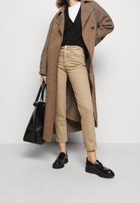 Abrigo largo de cuadros marrones, suéter negro, camisa blanca, jeans beige, zapatos negros, sosteniendo una bolsa tote negra; exhibición de un conjunto casual en capas.