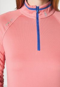 Růžové sportovní tričko s vysokým límcem a modrým zipem, s hladkým povrchem a reflexními logovými akcenty na hrudi.