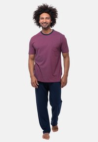 Glunderende man met krullend haar loopt op blote voeten, gekleed in een gestreept paarse t-shirt en donkerblauwe broek, tegen een effen witte achtergrond.