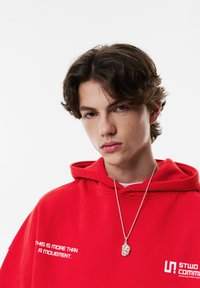 Punainen huppari, jossa etutasku ja valkoinen teksti "THIS IS MORE THAN A MOVEMENT." Hopeinen kaulakoru, jossa yksityiskohtainen riipus.