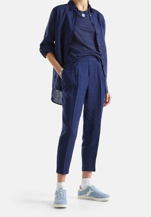 Personne portant une chemise boutonnée bleu marine sur un t-shirt bleu marine, un pantalon bleu marine assorti, des baskets bleu clair, des chaussettes blanches et des bijoux argentés, debout.