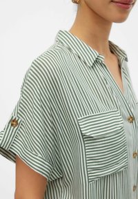 Roheline ja valge triibuline lühikeste varrukatega shirt, millel on krae, eesmine tasku ja pruunid nupud. Kangas on kerge ja sile.