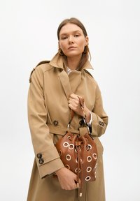 Brown leather bucket bag with large metallic eyelets, knotted closure, and braided handle. Featured with a beige trench coat.