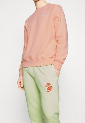 Osoba ubrana w blado różową bluzę Nike oraz jasnozielone spodnie dresowe Nike z trzema czerwonymi logo Nike na lewej nodze.