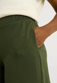 Pantalones verdes con cintura elástica, detalle de bolsillos y una tela texturizada. La tela parece ligera y ligeramente arrugada.