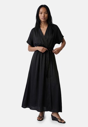 Donna che sistema la cintura in vita di un lungo abito nero a portafoglio con maniche corte, abbinato a sandali con zeppa neri, in piedi su sfondo bianco.