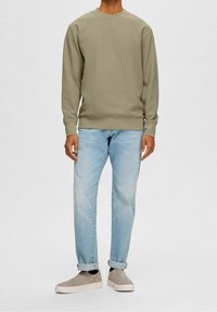 Světle olivová mikina s texturovaným povrchem, kombinovaná se světlými modrými džínami a šedými slip-on botami. Uvolněný střih s ohrnutými manžetami.