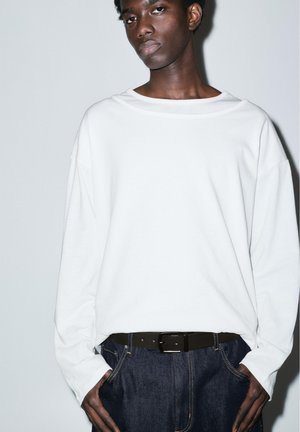 Tânăr cu pielea închisă la culoare, purtând o cămașă albă lejeră cu mâneci lungi, blugi albaștri închiși și o curea neagră, stând în picioare pe un fundal simplu.