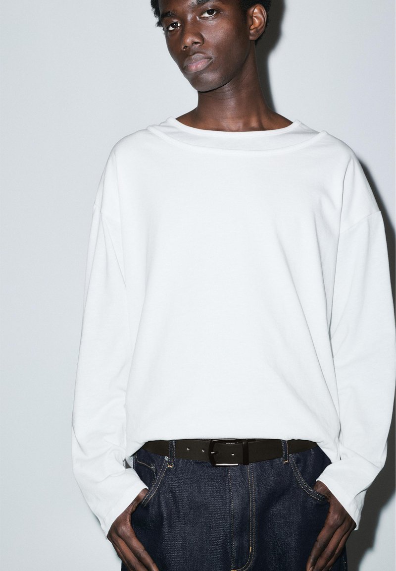 Jeune personne à la peau foncée portant une chemise blanche ample à manches longues, un jean bleu foncé et une ceinture noire, debout devant un fond uni.