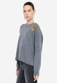 Maglione grigio a maglia con una vestibilità comoda, caratterizzato da un motivo decorativo giallo e oro sulla spalla sinistra e polsini a costine.