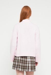 Gaiši rozā adīts džemperis ar augstu apkakli, ribotām aprocēm un apakšu, kombinēts ar brūnu rūtainu krokainu svārku. Mīksta tekstūra un ikdienišķa piegriezuma forma.