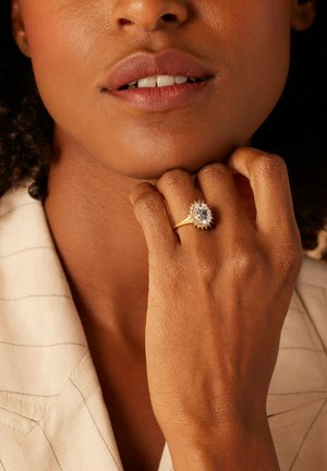 Goldring mit einem ovalen Kristall und einem strukturierten Band, getragen an einer Hand, die gegen das Kinn ruht, mit sichtbarer hellfarbener Kleidung.