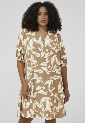 Femme aux cheveux bouclés portant une robe marron clair mi-genoux à imprimé floral blanc et manches bouffantes jusqu'aux coudes sur un fond uni.