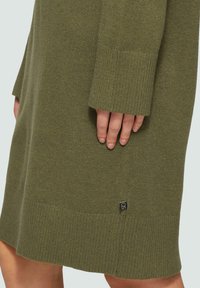 Grünes gestricktes Kleid mit geripptem Saum und Bündchen, versehen mit einem kleinen Logoschildchen nahe dem unteren Saum. Der strukturierte Stoff betont die Schlichtheit.