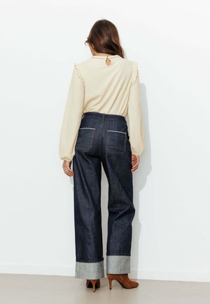 Blouse beige à manches longues avec épaules à volants et dos à ouverture en forme de goutte, associée à un jean large en denim foncé taille haute, avec ourlets retroussés.