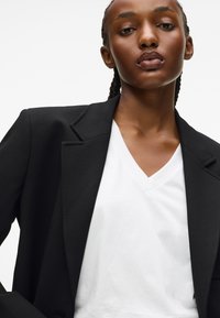 Čierny blejzer so štruktúrovanými ramenami a klopovými reverzmi, nosený cez biely tričko s výstrihom do v. Hladká tkanina s čistým, minimalistickým dizajnom.