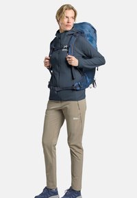 Sac à dos de randonnée bleu avec des bretelles, veste grise zippée avec capuche, pantalon beige clair, et chaussures de randonnée bleues. Matériaux texturés visibles.