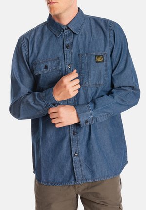 Chemise en denim bleu avec un design à boutons, deux poches poitrine et un logo en patch sur la poche gauche. Manches longues avec poignets à boutons.