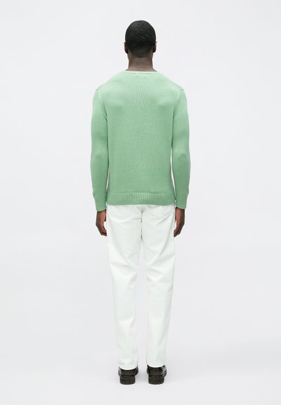 Zelena pletena majica z rebrastim robom, kombinirana z belimi hlačami z ravnimi nogami in črnimi čevlji, prikazana od zadaj na enostavnem ozadju.