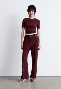 Burgundy fitted top with a white collar, paired with high-waisted trousers, accented by a black belt and brown shoes with white laces.