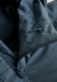 Prim-plan al unei jachete de culoare bleumarin, arătând un fermoar și un șnur ajustabil cu inele de plastic și opritor pe glugă.