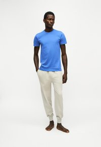 Un homme est debout pieds nus, portant une chemise bleue à manches courtes et un pantalon de jogging beige clair sur un fond blanc uni.