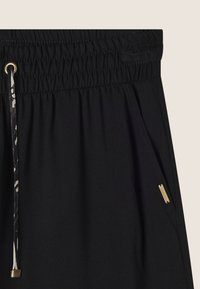 Pantaloni neri con elastiche in vita e dettagli in hardware dorato, con nastrino e tasche laterali, realizzati in un tessuto morbido e leggermente strutturato.