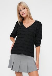 Zwarte blouse met textuur, een V-hals en driekwartmouwen, voorzien van horizontale strepen. Gecombineerd met een grijze geplooide rok.