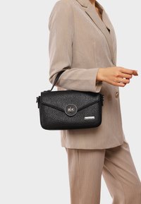 Borsa a tracolla in pelle nera con texture, chiusura a pattina, hardware circolare in argento e tracolla removibile, indossata con un completo beige.