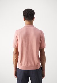Uomo con capelli ricci e corti che indossa una maglietta a maniche corte di colore rosa e pantaloni scuri, in piedi rivolto di spalle su uno sfondo bianco.
