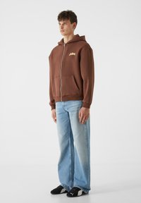 Rudas užtrauktukas su kišene priekyje, su auksiniu logotipu. Dėvimi su šviesiai mėlynomis plačiomis kelnėmis ir juodais sportiniais bateliais.