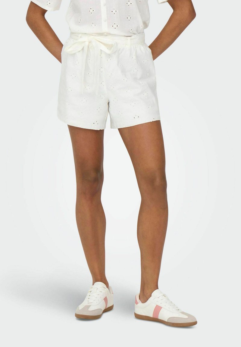Pantalones cortos de algodón blanco con patrón de ojales, cintura con cordón y corte relajado, combinados con zapatillas blancas con rayas rosas y acentos beiges.