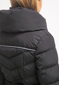 Chaqueta de plumas negra con un alto cuello, diseño acolchado y un acento reflectante a lo largo de la costura trasera. Textura suave y lisa.