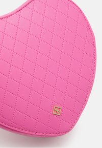 Růžová kosmetická taštička s prošívaným vzorem, hladkým povrchem, ve tvaru srdce a zlatým logem na jedné straně.