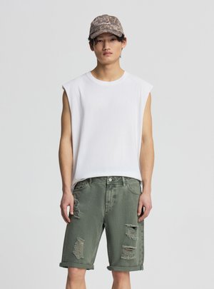 Młody mężczyzna w białej koszulce bez rękawów, zniszczonych zielonych szortach jeansowych i moro czapce z daszkiem z napisem „Las Vegas".