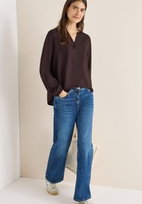 Tmavě hnědá blůza s dlouhým rukávem a výstřihem ve stylu henley, doplněná černými knoflíky, v kombinaci s světle modrými širokými kalhotami a bílými teniskami.