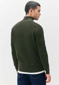 Hombre de espaldas vistiendo un pulóver de punto verde oscuro texturizado con cuello alto y jeans azul oscuro, frente a un fondo blanco liso.