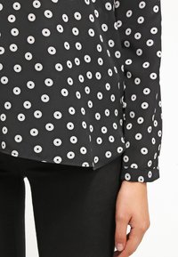 Blusa nera con motivi circolari bianchi, maniche lunghe e orlo curvo. Tessuto liscio con leggero lucido e polsini con bottoni.