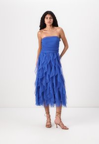 Strapless blue dress with layered tulle ruffles, fitted bodice, and ankle-length hem. Paired with strappy beige heels.