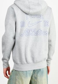 Felpa grigia con cappuccio regolabile. Presenta la scritta ricamata in viola "Nike Athletics 1972" sul retro. Materiale morbido in misto cotone.