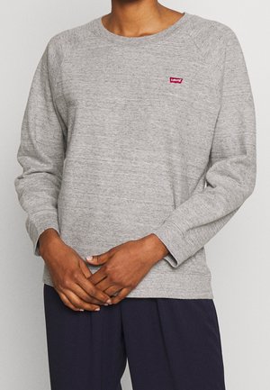 Person wearing a light grey long-sleeve Levi’s sweatshirt and dark blue pants with hands gently clasped at waist.