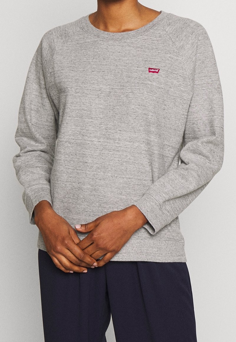 Personne portant un sweat-shirt Levi’s à manches longues gris clair et un pantalon bleu foncé, les mains doucement jointes à la taille.