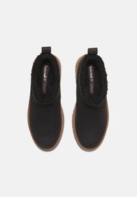 Black slip-on shoes with a textured black fleece collar and tan rubber sole; smooth leather upper with minimal branding.